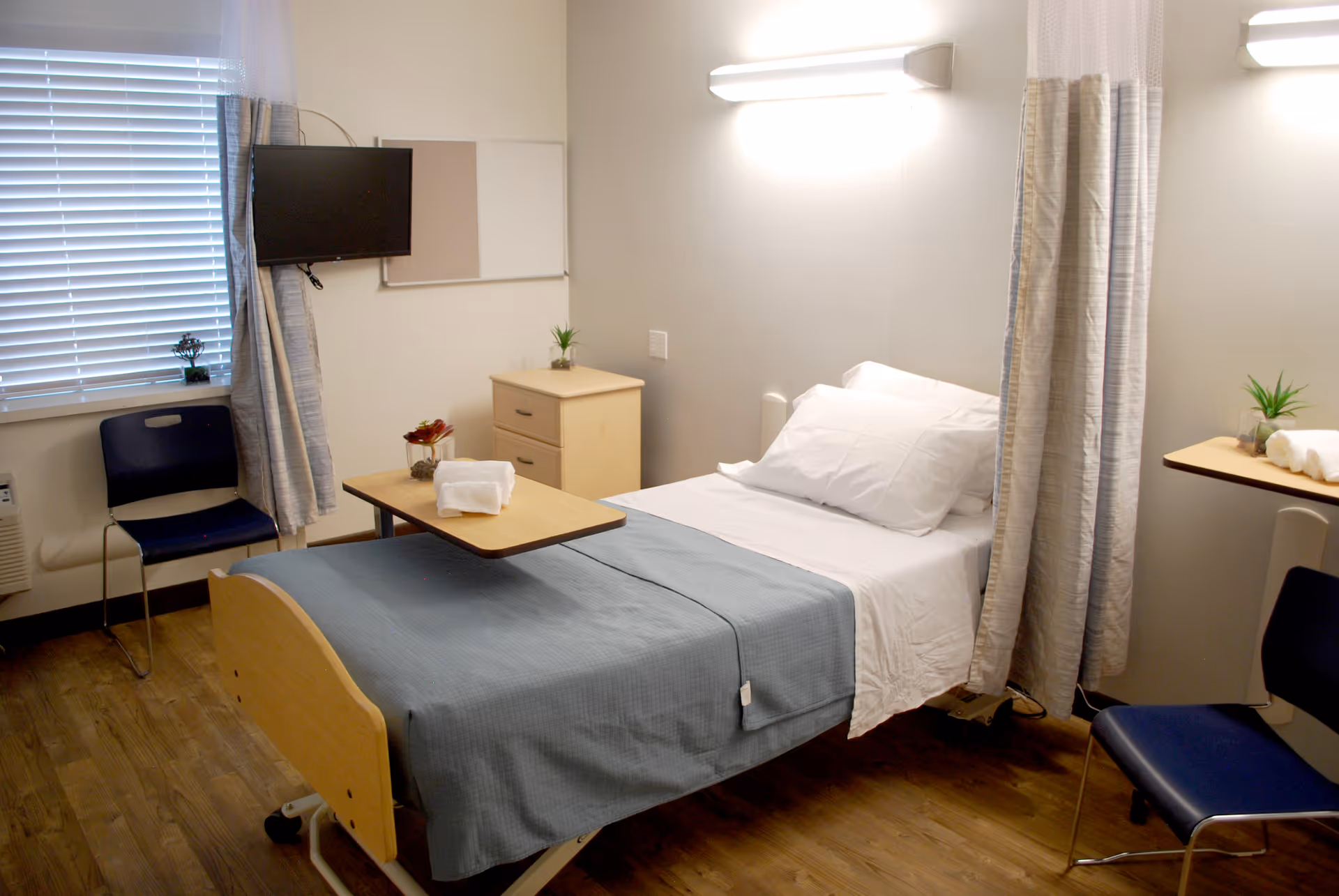 A clean and simple bedroom in a senior living facility with a single bed covered with a blue blanket and white pillows. There is a small wooden bedside table with a plant and a towel on a movable overbed table. Two blue chairs are placed near the window with blinds and a wall-mounted TV. The room has wooden flooring and light-colored walls with a curtain partially drawn around the bed.