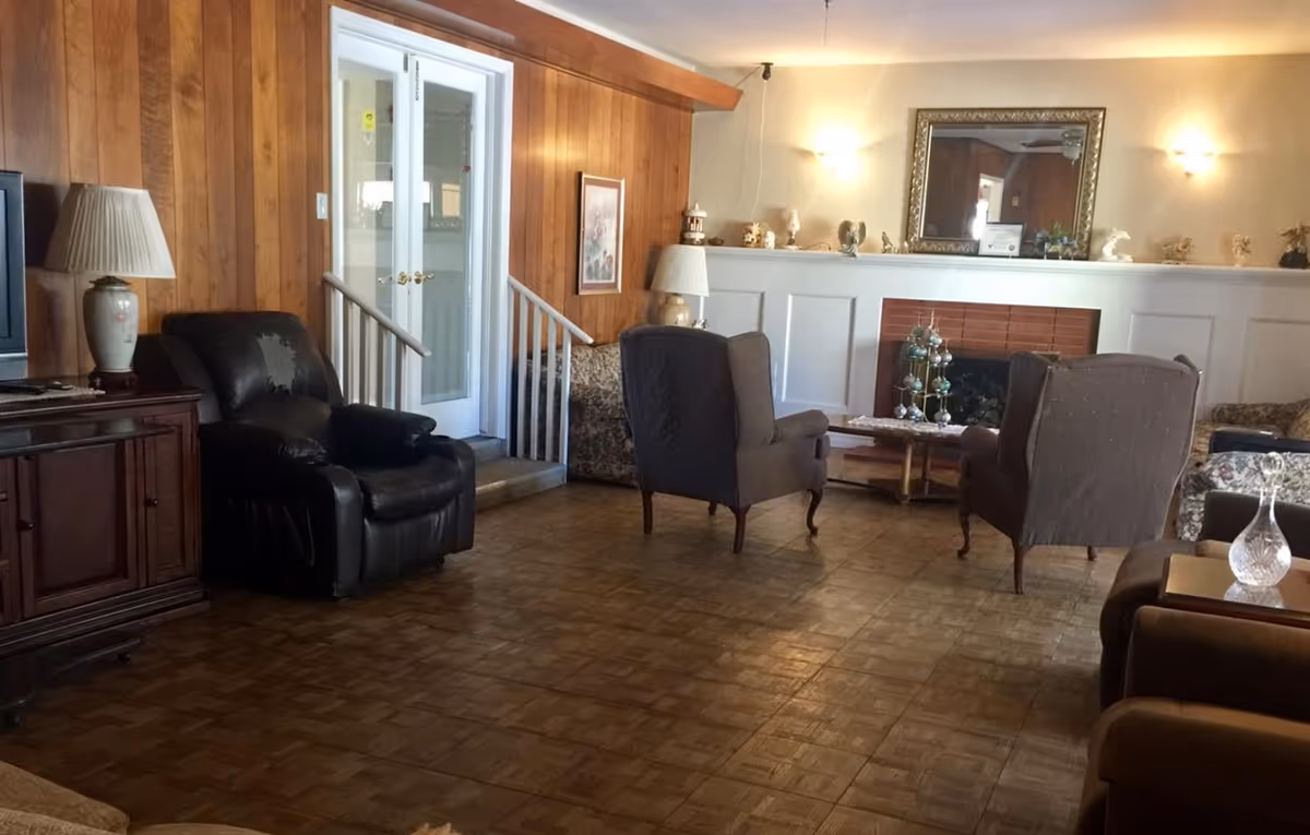 A cozy living room with wooden paneled walls, a parquet floor, and a white fireplace with decorative items on the mantel. The room contains several armchairs, a floral sofa, a wooden cabinet with a lamp, and a large mirror above the fireplace. French doors with glass panels are visible in the background.