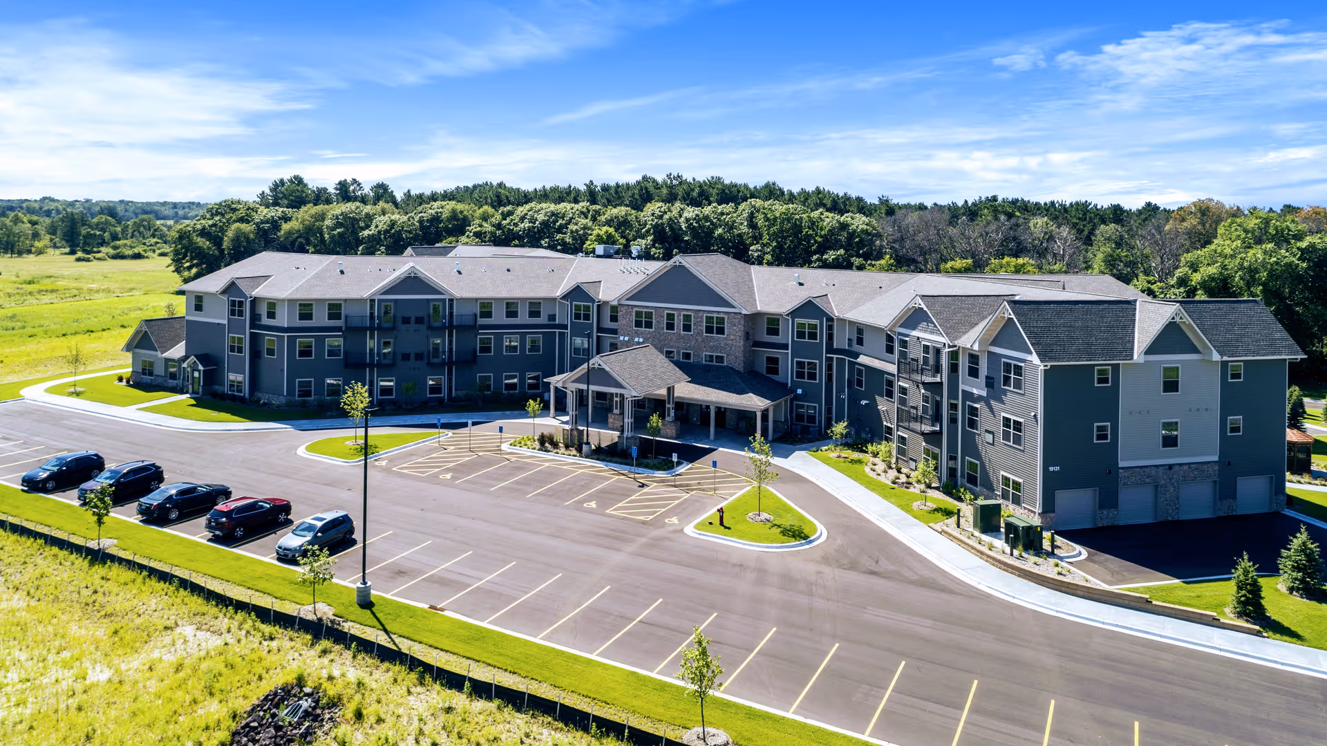 Aerial view of a three-story senior living building with a covered main entrance, adjacent parking lot, and surrounding trees and grassy fields.