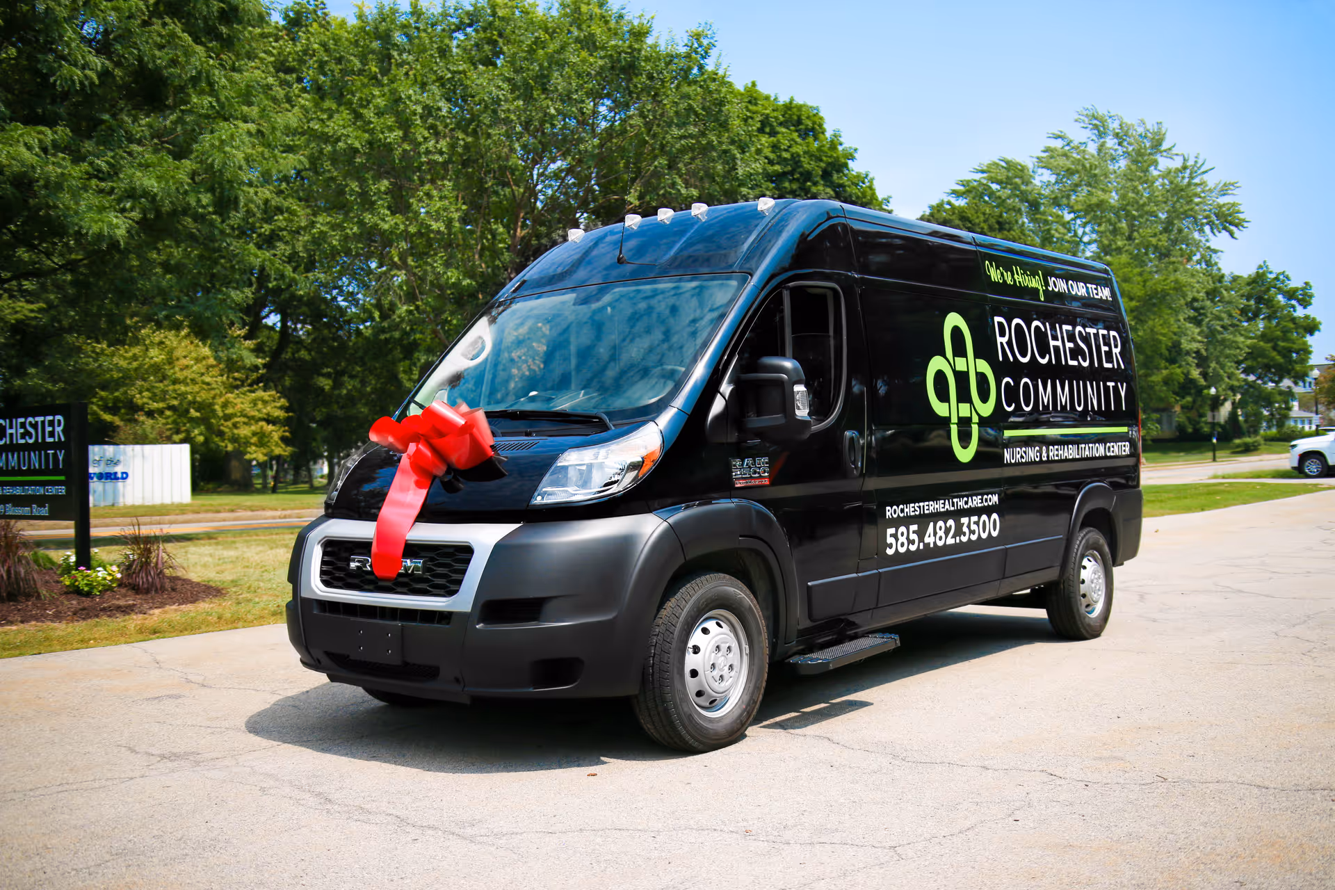A black Rochester Community Nursing & Rehabilitation Center van parked on a paved area with a large red bow on its front grille. The van features the center's logo, contact number, and website on its side. The background includes green trees and a clear blue sky.