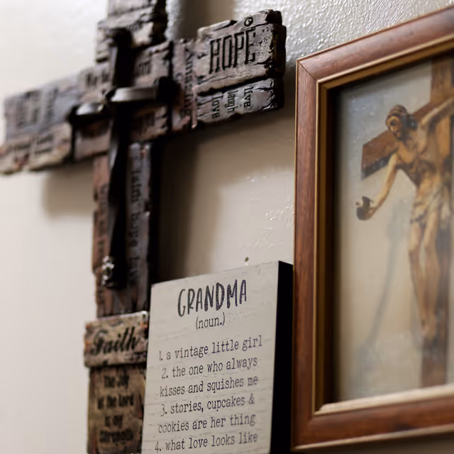 Close-up view of a wall decorated with a wooden cross featuring words like 'HOPE' and 'Faith', a framed picture of Jesus on the cross, and a wooden plaque defining 'GRANDMA' with affectionate descriptions.