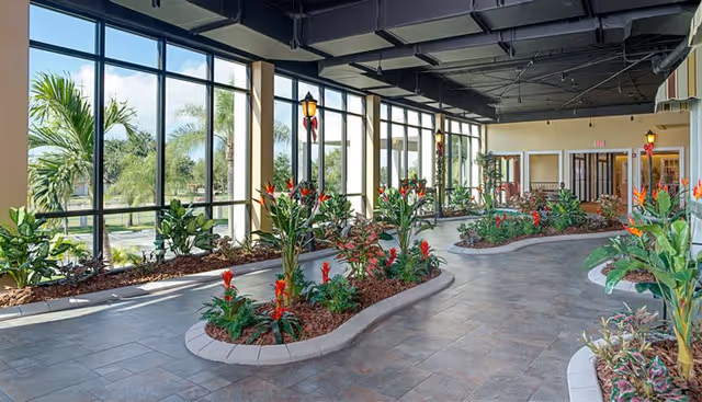 Indoor garden area with large windows allowing natural light to flood the space. The garden beds contain various green plants and red flowers, and the floor is tiled. The ceiling is exposed with visible ducts and lighting fixtures. There are doors and seating areas visible in the background.
