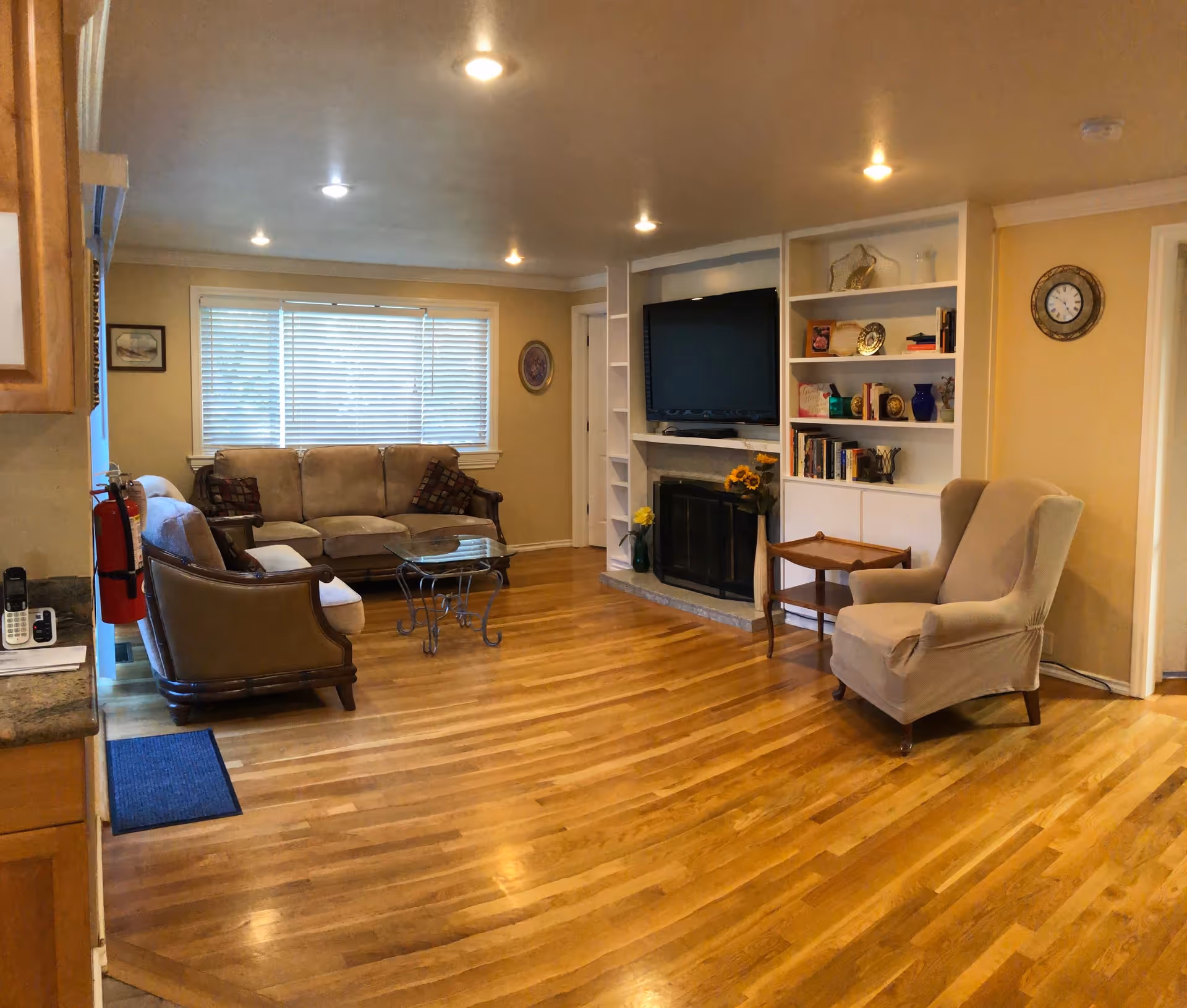 A cozy living room with wooden floors, a beige sofa set with patterned cushions, a glass coffee table with a metal frame, a beige armchair, a wall-mounted TV above a fireplace, built-in white shelves with books and decorative items, a clock on the wall, and a window with blinds letting in natural light.
