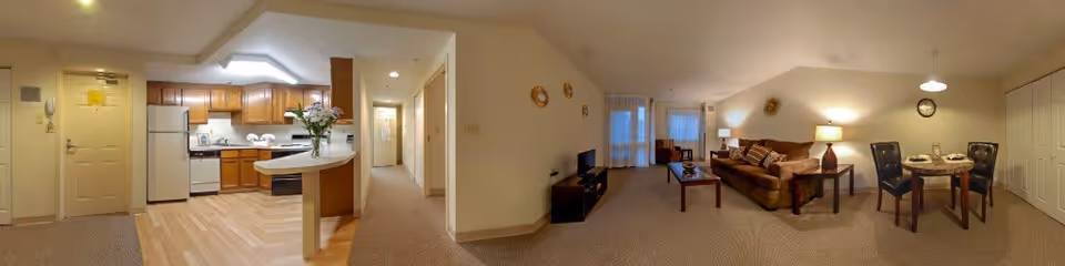 Wide panoramic view of an apartment interior showing an open kitchen with wooden cabinets, white refrigerator, stove, and a small breakfast bar with flowers on it. Adjacent to the kitchen is a carpeted hallway leading to other rooms. The living area features a brown sofa, coffee table, side table with a lamp, a TV on a stand, and a small dining table with four chairs. The room is softly lit with ceiling and table lamps, and there are windows with curtains in the background.