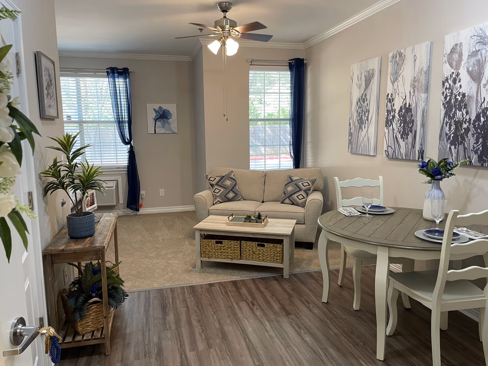 A cozy living and dining area in an apartment with a beige loveseat sofa, two patterned cushions, a wooden coffee table with baskets underneath, and a round dining table set for two with white chairs. The room has wood flooring in the dining area and carpet in the living area, with two windows covered by blue curtains. There are three floral paintings on the wall above the dining table, a ceiling fan with lights, and a wooden console table with potted plants near the entrance.