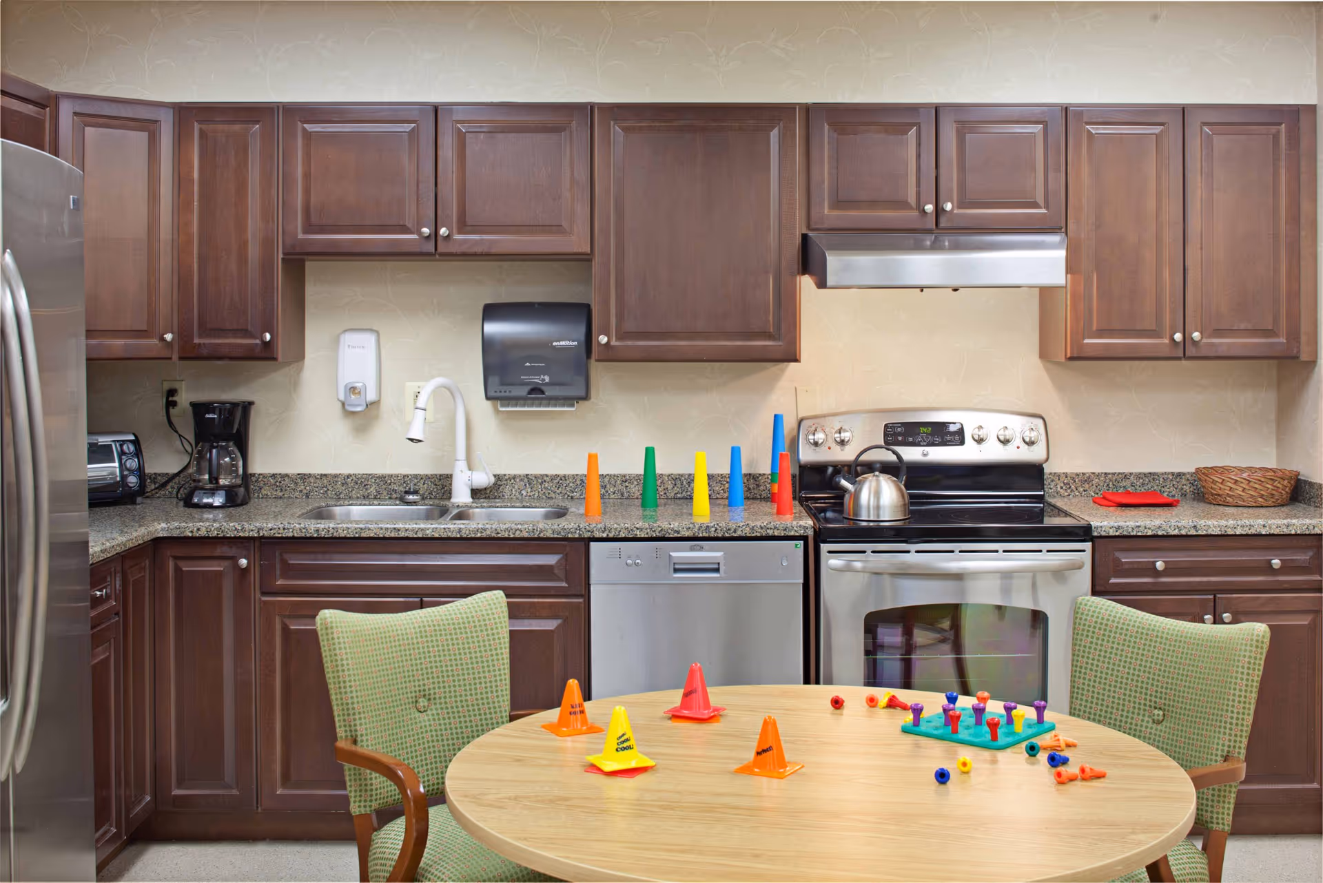 A kitchen area with dark wooden cabinets, a stainless steel refrigerator, dishwasher, and stove. On the countertop, there are colorful plastic cones and a kettle on the stove. In front of the kitchen is a round wooden table with two green cushioned chairs, and the table has more colorful cones and small plastic pieces scattered on it.