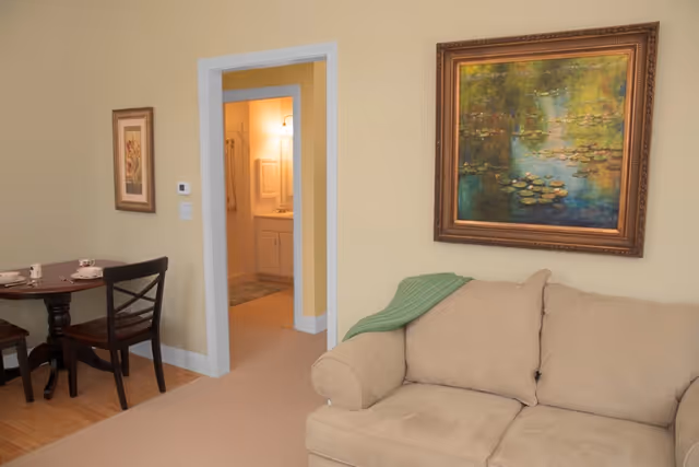 A cozy living area with a beige couch draped with a green blanket, a framed painting of a pond with lily pads on the wall above the couch, and a small round wooden dining table with two chairs. An open doorway leads to a bathroom with a lit vanity mirror.