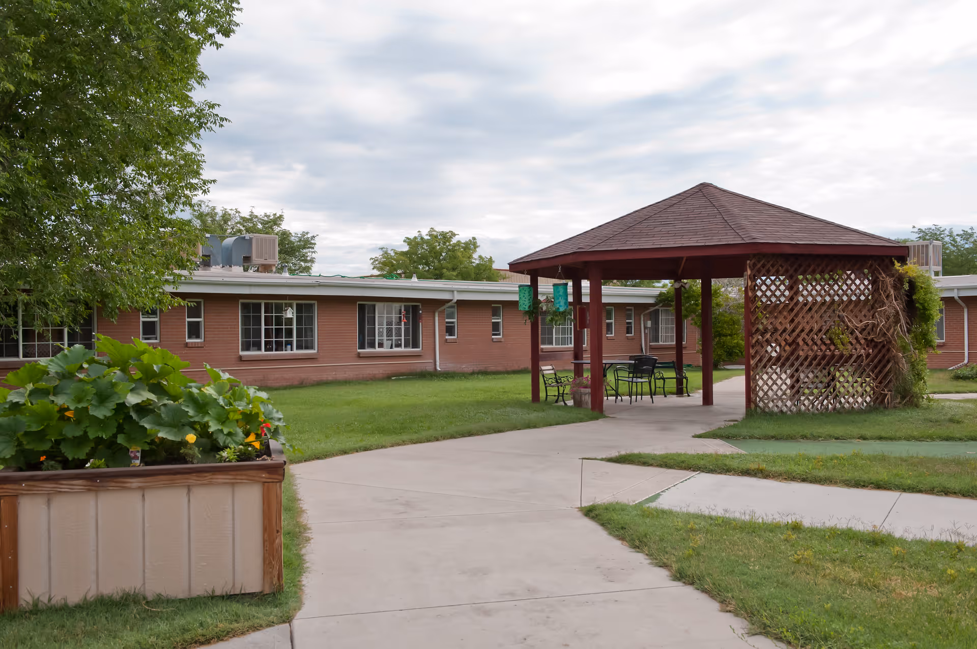 Outdoor courtyard area of a senior living facility with a paved walkway, a wooden gazebo with lattice sides, outdoor chairs and tables, green grass, and a planter box with leafy plants. The building in the background is a single-story brick structure with multiple windows. The sky is cloudy.