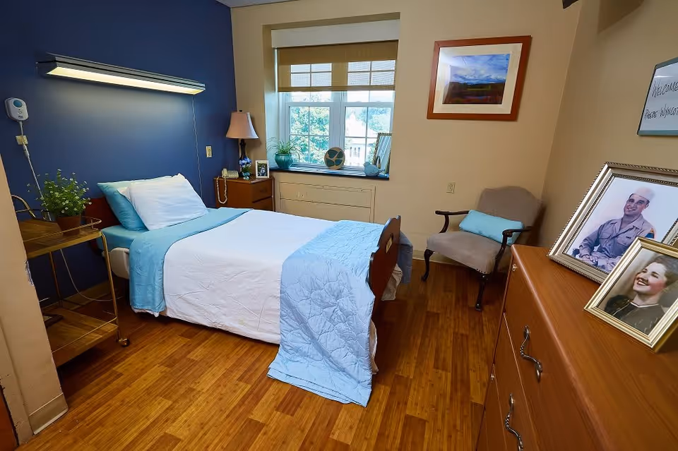 A tidy senior bedroom with a single bed dressed in light blue bedding, a window, armchair, dresser with framed photos, and wood flooring.