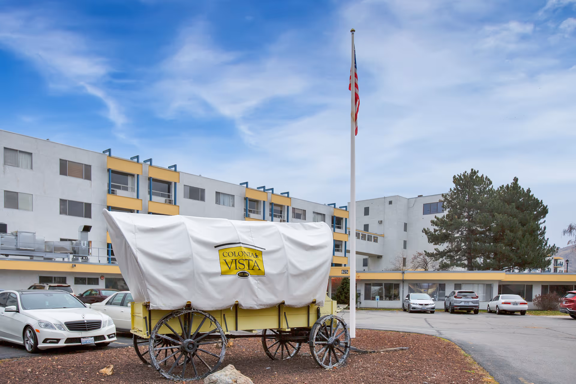 Exterior view of Colonial Vista Post Acute facility showing a covered wagon with the facility's name on it, an American flag on a flagpole, several parked cars, and a multi-story building with yellow accents under a partly cloudy sky.