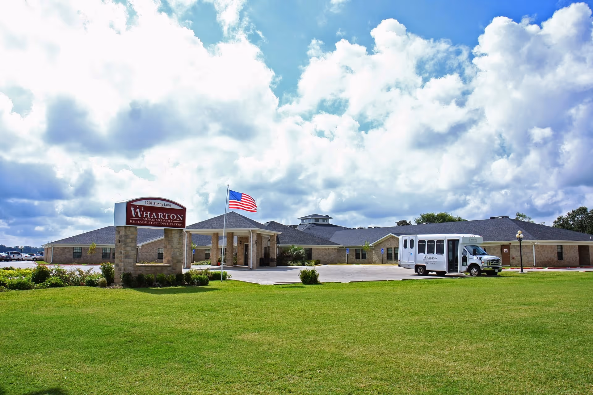 Exterior view of the Wharton Nursing and Rehabilitation Center building with a large lawn, an American flag, and a parked shuttle bus near the entrance.