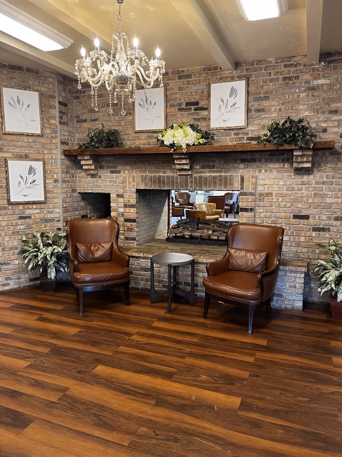 Cozy lounge with two brown leather armchairs and a small round table in front of a brick fireplace under a chandelier.