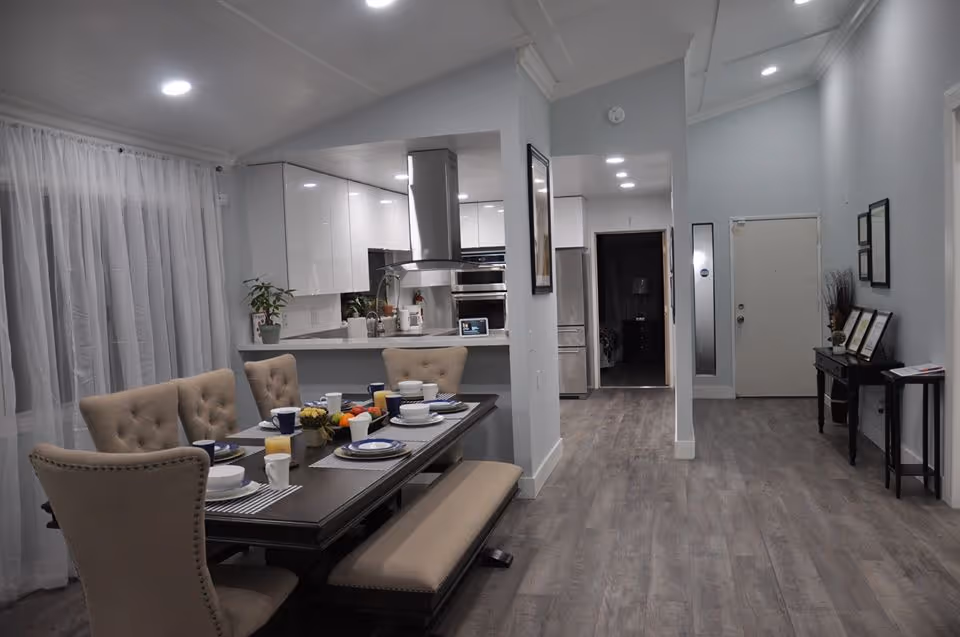 A modern dining area with a dark wooden table set for six, including plates, cups, and glasses with orange juice. The dining chairs are upholstered in beige fabric, and there is a matching bench on one side of the table. Behind the dining area is a kitchen with white cabinets, stainless steel appliances, and a range hood. The floor is covered with wood-like vinyl planks, and the walls are painted light gray. To the right, there is a hallway leading to a door, with a console table and framed pictures on the wall.