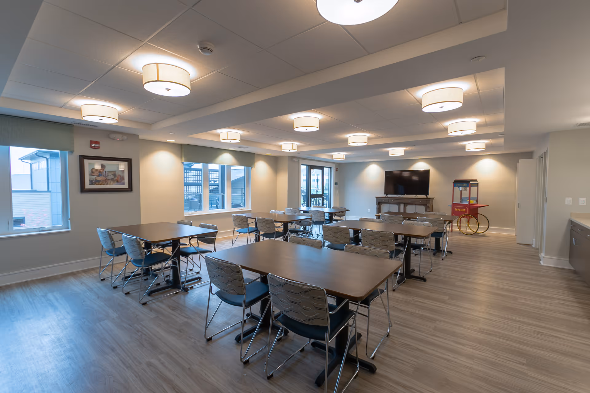 A spacious, well-lit dining room with multiple rectangular tables and chairs arranged neatly. The room features large windows with green valances, modern ceiling lights, a flat-screen TV on a wooden stand, and a vintage-style popcorn machine in the corner. The flooring is light wood, and the walls are painted a soft neutral color.