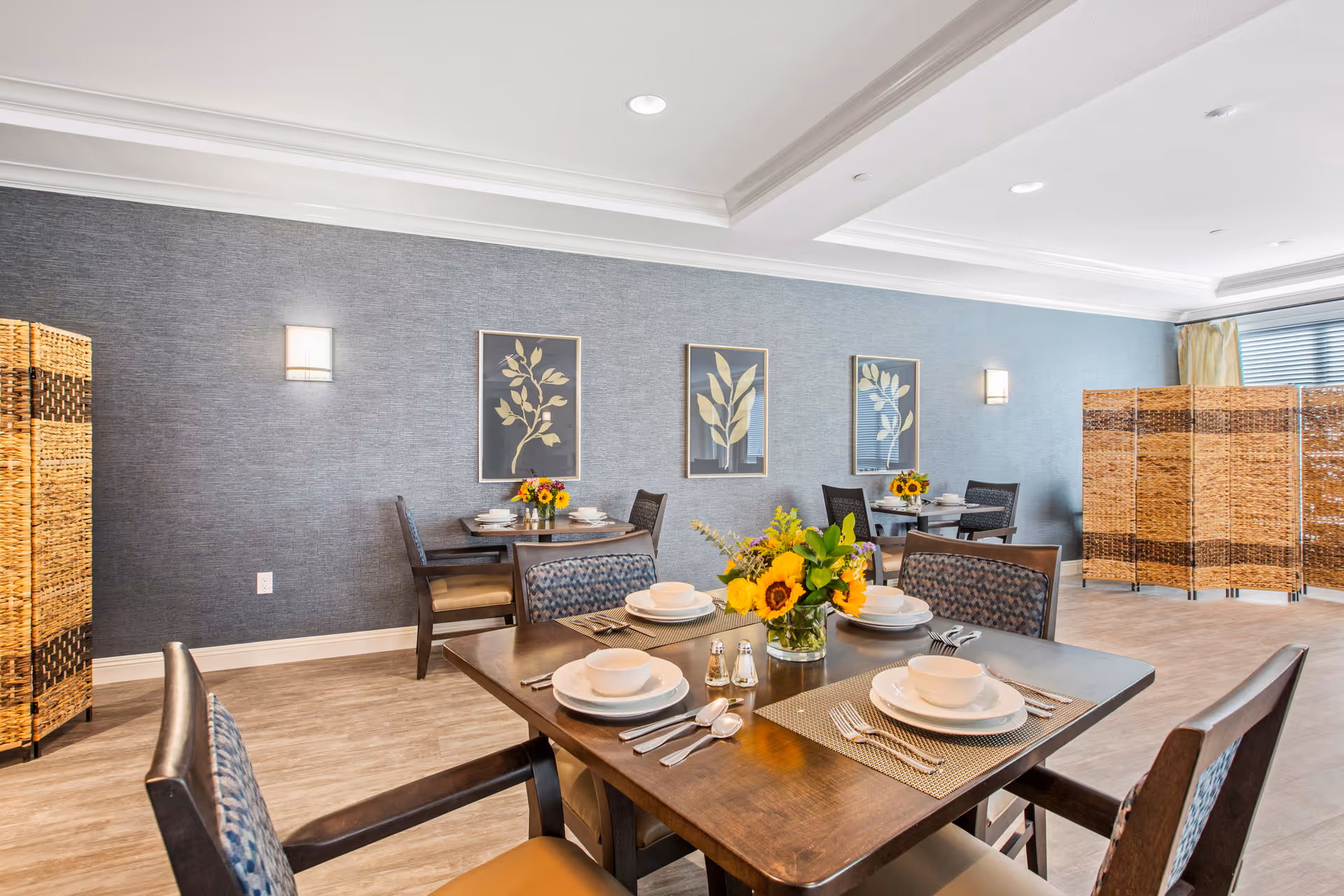 A bright dining room with wooden tables set with white plates, bowls, and silverware. Each table has a centerpiece with sunflowers and other flowers. The walls are painted blue with three framed botanical prints and two wall sconces. There are woven room dividers on the right side near large windows with blinds.