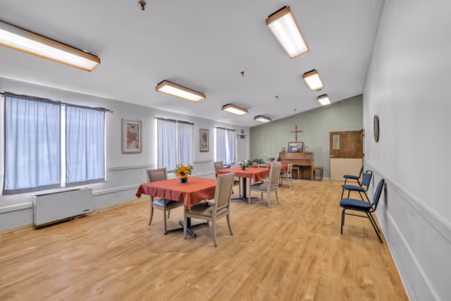 A bright communal dining room with tables covered in red tablecloths, chairs, a piano against the far wall, and a cross hanging above it.
