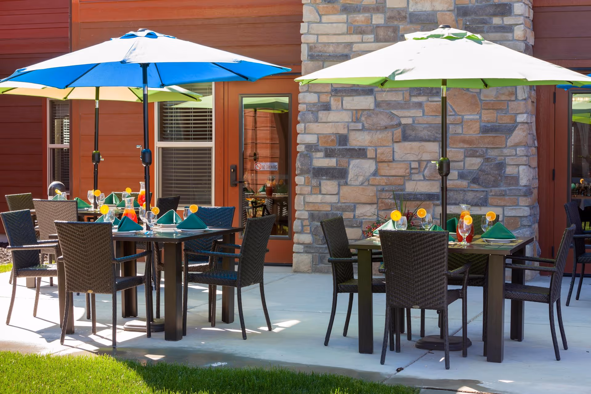 Outdoor patio dining area with tables, wicker chairs, colorful umbrellas, and place settings outside a stone and red-sided building.