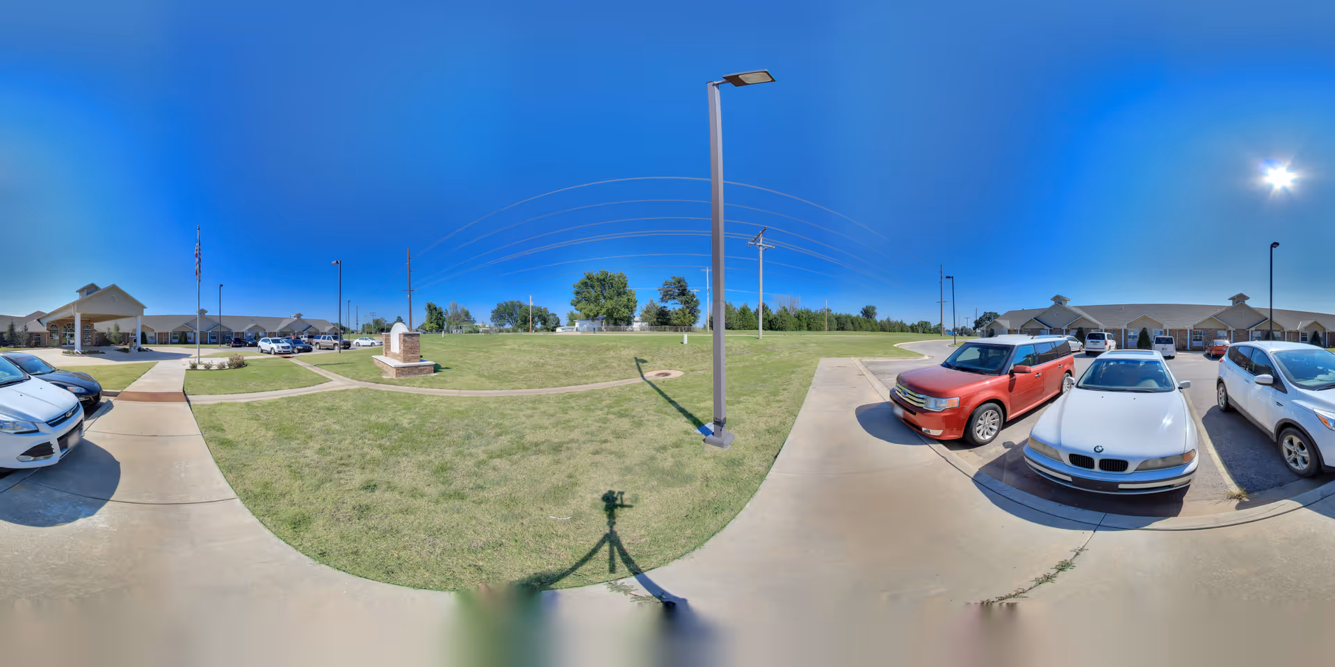Front exterior of a senior living facility with parked cars, a grassy courtyard, walkways and lamp posts under a clear blue sky.