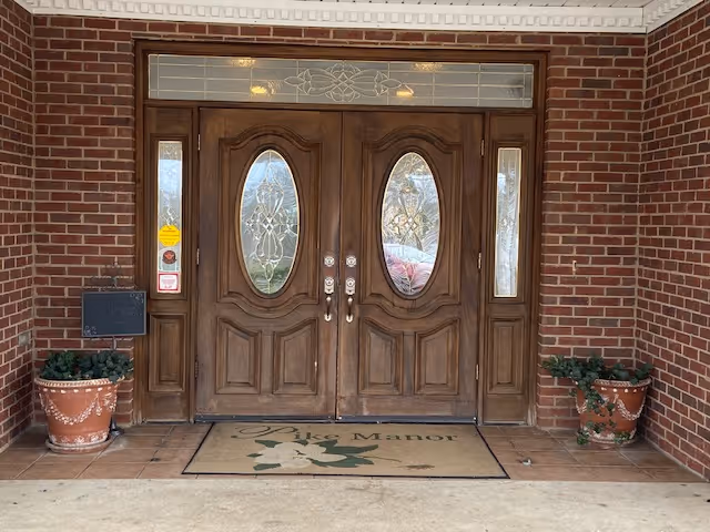 Double wooden decorative front doors with oval glass panels in a brick entryway, flanked by potted plants and a doormat reading 'Pike Manor'.
