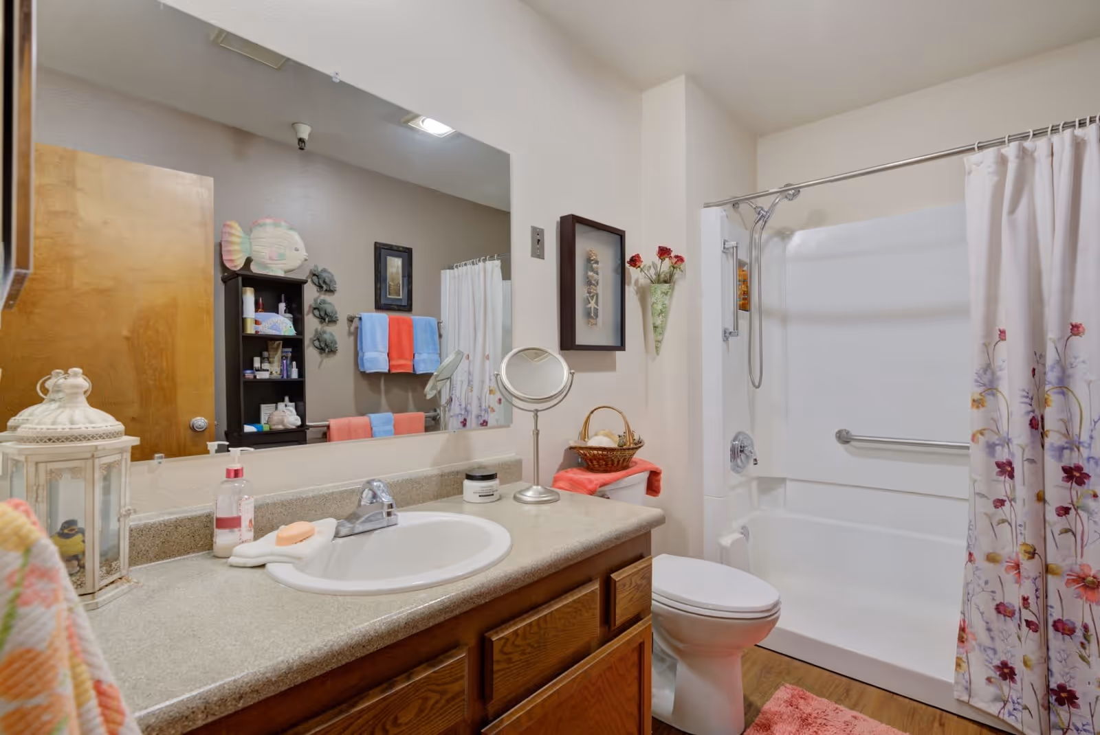A clean and well-lit bathroom featuring a countertop with a sink, a large mirror, and various toiletries. There is a toilet next to a bathtub with a floral shower curtain. The bathroom has wooden cabinets, a towel rack with colorful towels, and decorative items including a basket with flowers and wall art.
