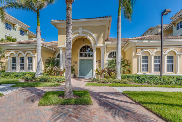 Front exterior view of a Mediterranean-style senior living facility building with a light yellow facade, arched windows, a central entrance with a green door, palm trees, and a paved walkway under a clear blue sky.