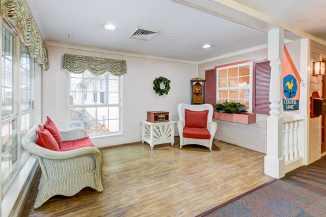 A cozy sitting area with two white wicker chairs and a matching loveseat, all with red cushions. There is a small white side table with a decorative box on it, a green wreath on the wall, and a window with pink shutters and a flower box beneath it. The floor is wooden, and large windows with floral valances let in natural light.