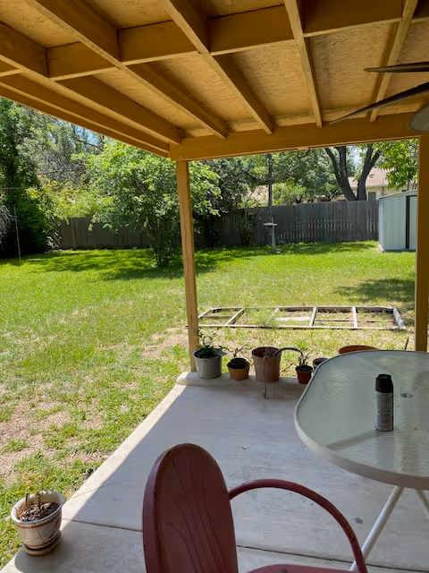 Covered patio with a table and chair overlooking a grassy backyard with potted plants, a fence, and a small shed.