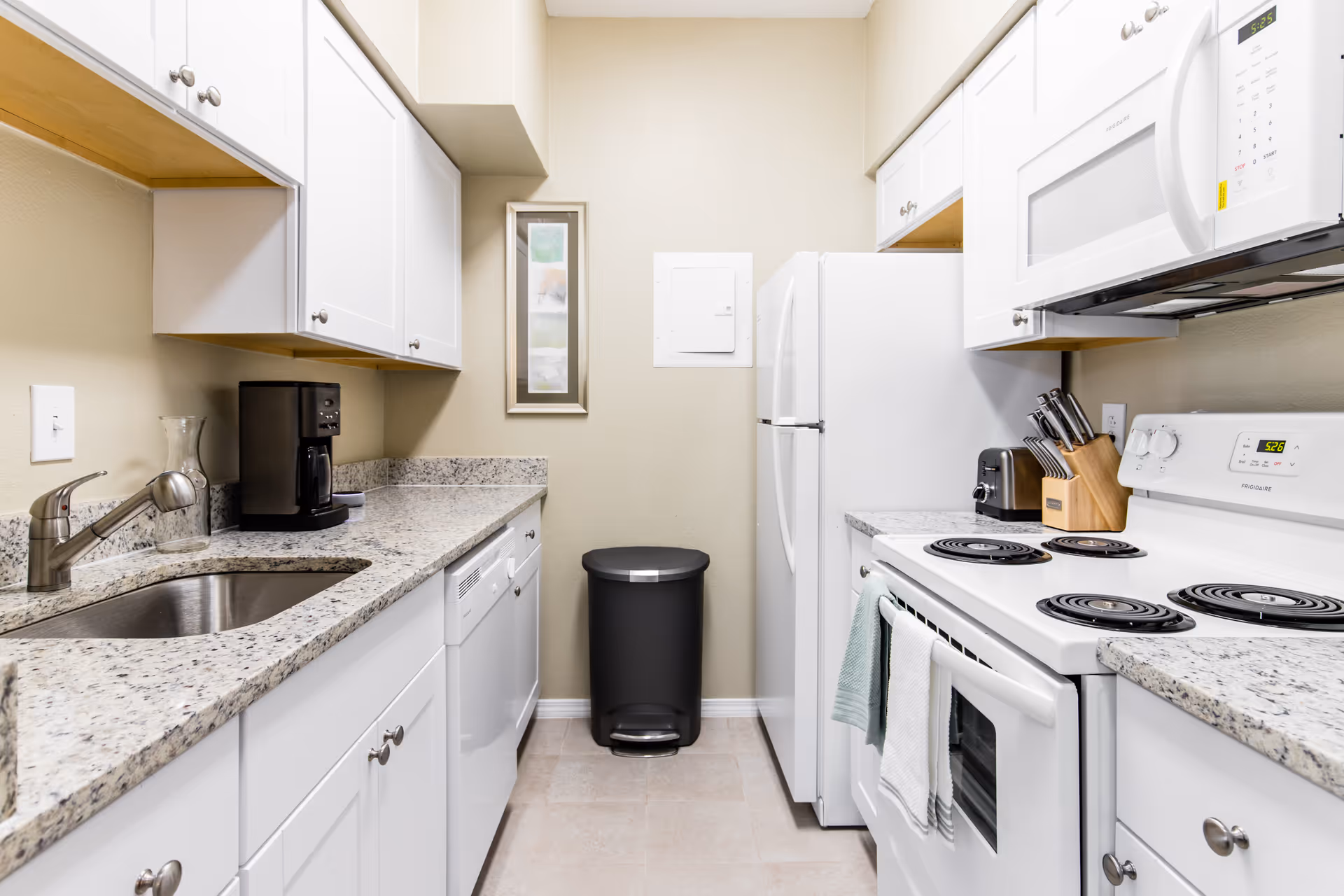 A clean, modern kitchen with white cabinets and granite countertops. The kitchen features a stainless steel sink, a black coffee maker, a dishwasher, a white refrigerator, a white electric stove with four coil burners, a white microwave above the stove, a knife block, a toaster, and a black trash can against a beige wall with a framed picture.