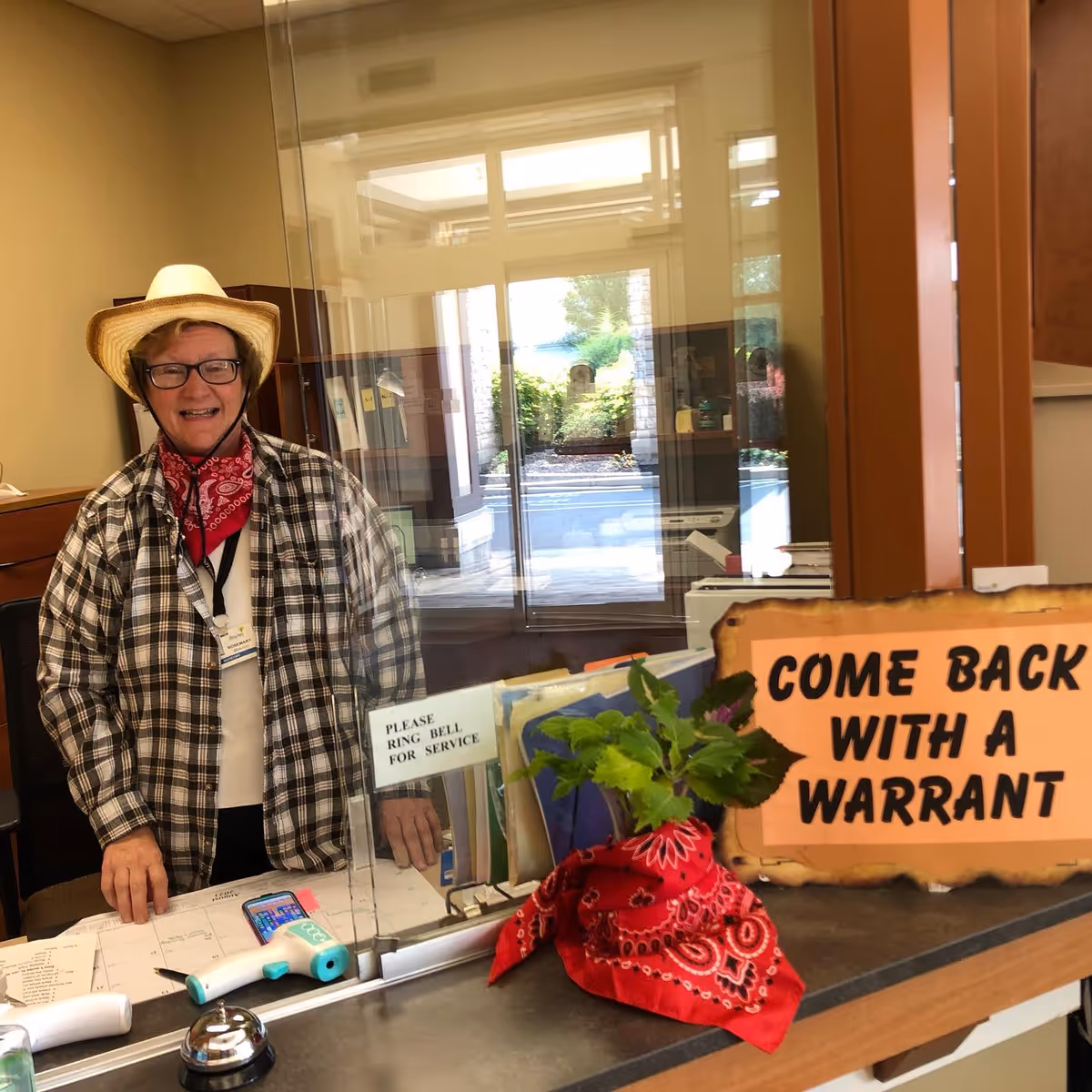 A smiling elderly woman wearing a straw hat, glasses, a red bandana around her neck, and a plaid shirt stands behind a glass partition at a reception desk. On the desk, there is a sign that says 'PLEASE RING BELL FOR SERVICE,' a small bell, a thermometer, a phone, and a decorative plant with a red bandana around its pot. Another sign on the desk reads 'COME BACK WITH A WARRANT.' The background shows a bright entrance area with glass doors and windows.