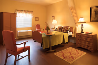 A cozy bedroom in a senior living facility with a single bed covered with a green blanket and striped pillows. There is a wooden bedside table with a lamp and books, a small rug on the wooden floor, two comfortable chairs, a window with a floral valance, and a wooden wardrobe. The room is warmly lit with soft yellow lighting.