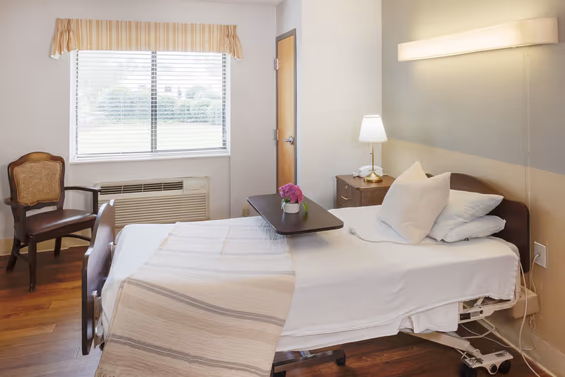 A single hospital-style bed with white linens and pillows in a well-lit room. There is a small wooden table on the bed with a pink flower in a white pot. To the right of the bed is a nightstand with a lamp and a telephone. A wooden chair is placed near a window with blinds and a beige valance. The floor is wooden, and the walls are painted in neutral tones.