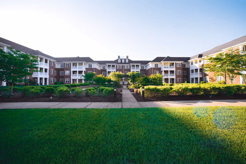 Exterior view of a large, multi-story senior living facility building with brick and white siding, surrounded by well-maintained green lawns, shrubs, and trees under a clear sky.