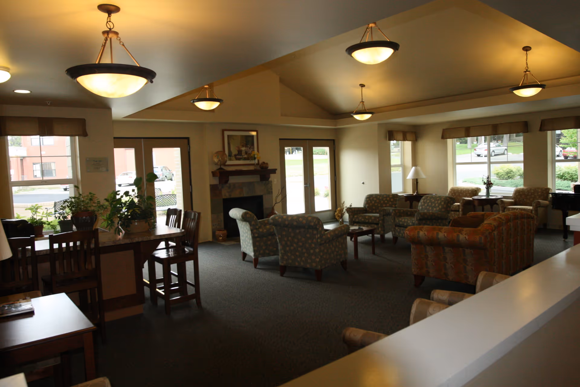 A spacious senior living facility common area with multiple armchairs and sofas arranged around small tables. The room features large windows letting in natural light, a fireplace with a framed picture above it, and several ceiling lights. There are also some plants on a counter with wooden chairs nearby.