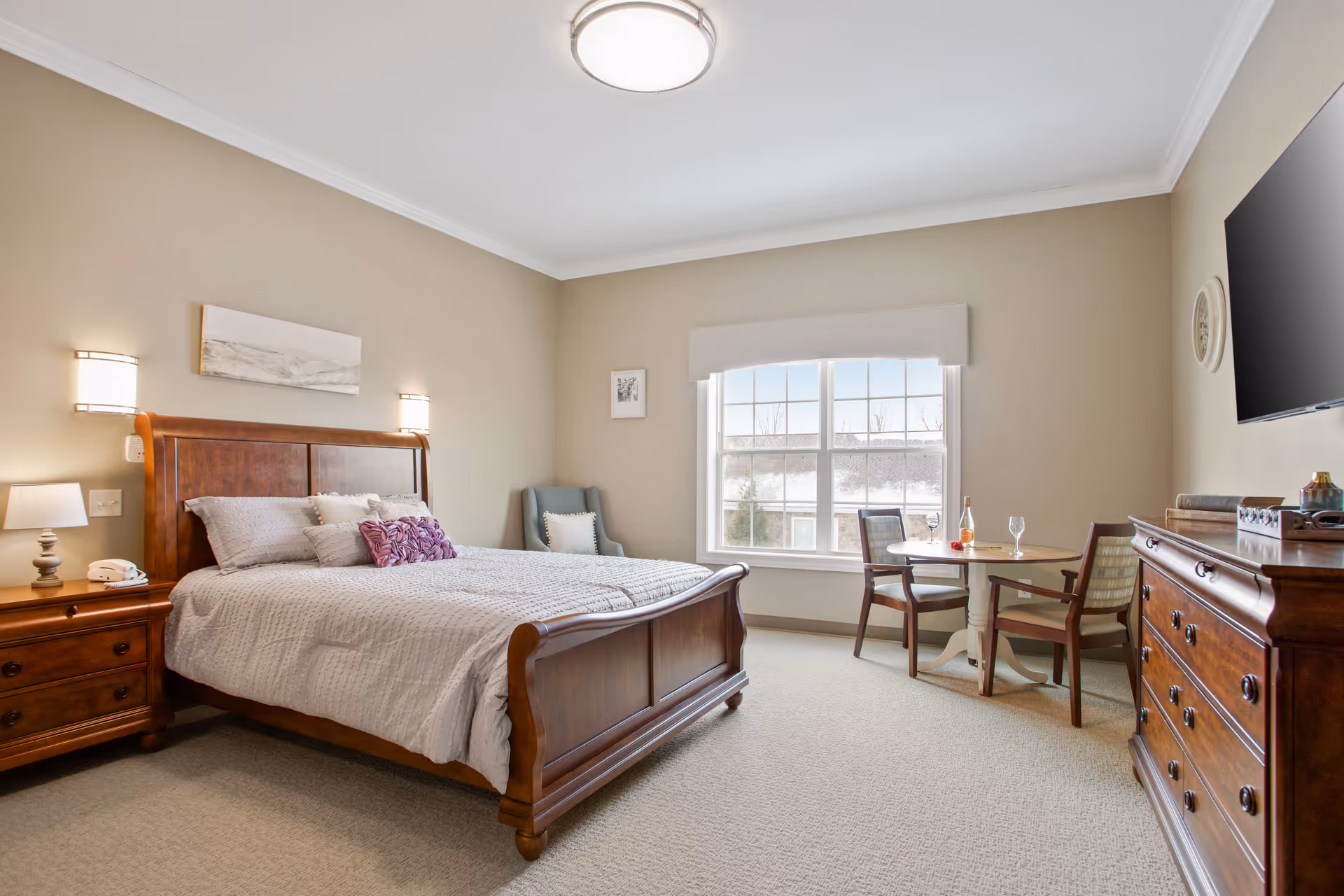 A well-lit bedroom in a senior living facility featuring a wooden bed with gray bedding and decorative pillows, a wooden nightstand with a lamp and phone, a comfortable armchair in the corner, a round table with two chairs near a large window, and a wooden dresser with a flat-screen TV mounted above it.