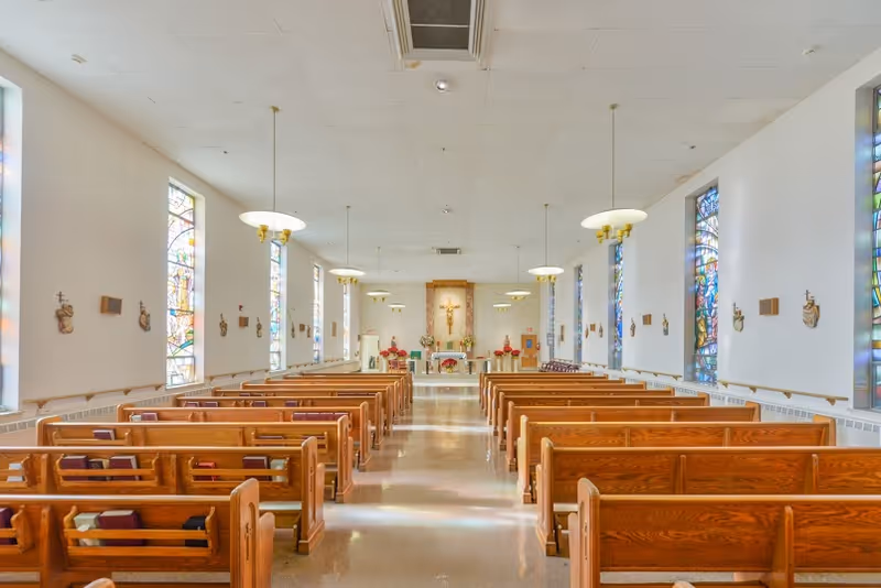 Interior view of a chapel with wooden pews arranged in rows facing an altar. The walls have stained glass windows and religious decorations. The ceiling has hanging lights and an air vent.