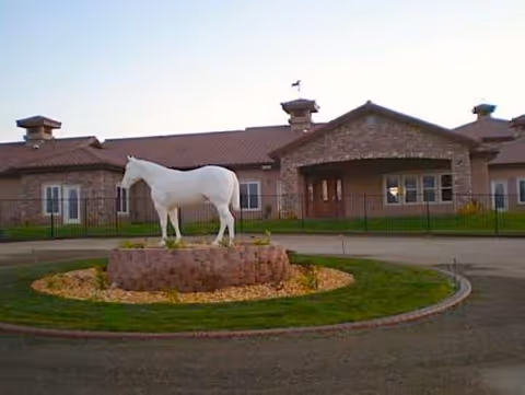 Front exterior view of a single-story brick building with a brown roof, featuring a circular driveway with a landscaped island in the center that has a white horse statue on a stone pedestal.