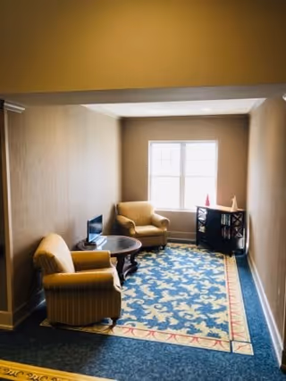 A small sitting area with two beige armchairs and a round wooden table between them, placed on a blue patterned rug. There is a window at the far end letting in natural light, and a dark wooden cabinet with decorative items on top is positioned against the wall beneath the window. The walls are painted beige.