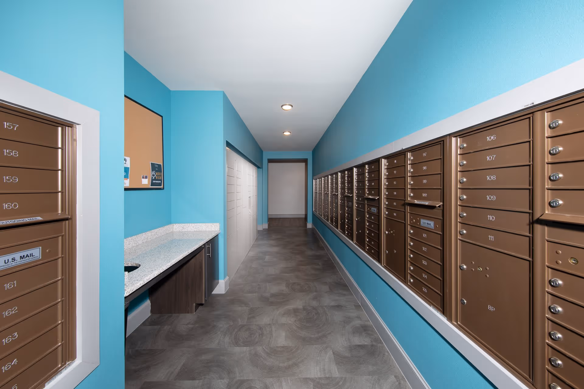 A hallway with bright turquoise walls and gray patterned flooring featuring a row of brown mailboxes on the right wall and a granite countertop with a small sink on the left side. The hallway is well-lit with ceiling lights and has a bulletin board on the left wall.