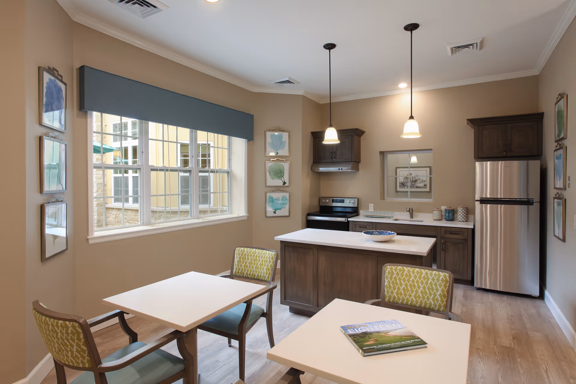 A modern kitchen and dining area with two square tables and four chairs featuring green patterned upholstery. The kitchen has a central island, stainless steel refrigerator, stove, and wooden cabinets. The room is well-lit with two pendant lights and a large window with a blue valance. Decorative framed artwork is displayed on the walls.