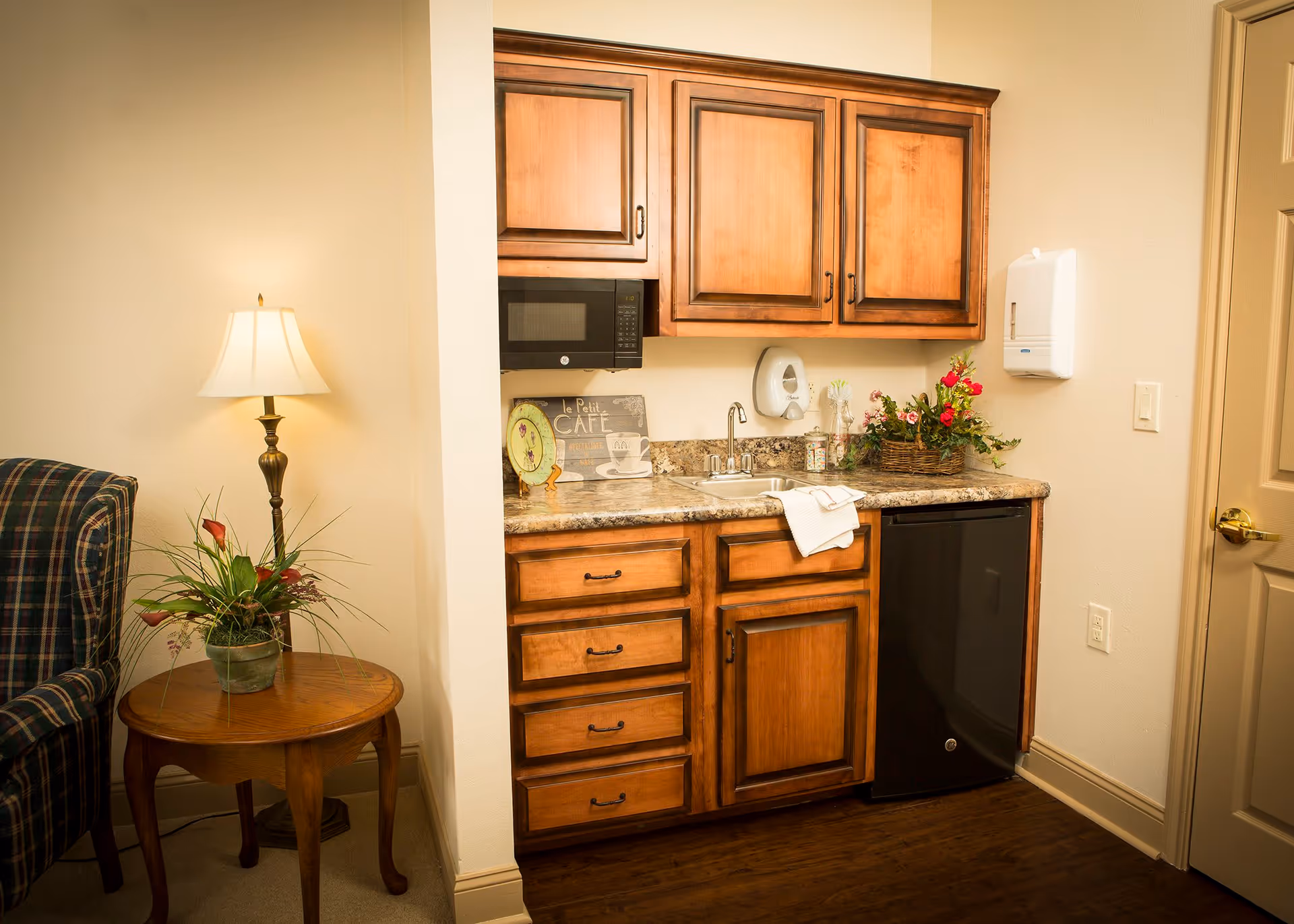 A small kitchenette area with wooden cabinets, a granite countertop, a sink, a black mini refrigerator, and a microwave mounted above. There is a decorative sign, a soap dispenser, and a flower arrangement on the counter. To the left, there is a wooden side table with a potted plant and a floor lamp next to a plaid upholstered chair.
