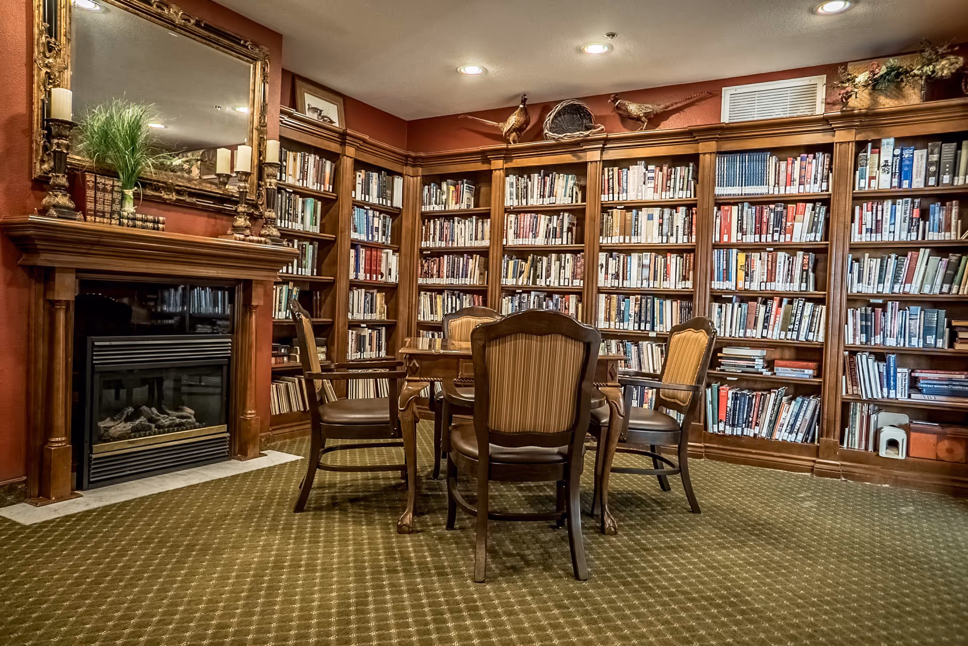 A cozy library room with wooden bookshelves filled with books lining the walls. In the center, there is a wooden table surrounded by four upholstered chairs. On the left side, there is a fireplace with a decorative mirror and candles on the mantel. The room has warm lighting and a patterned carpet.