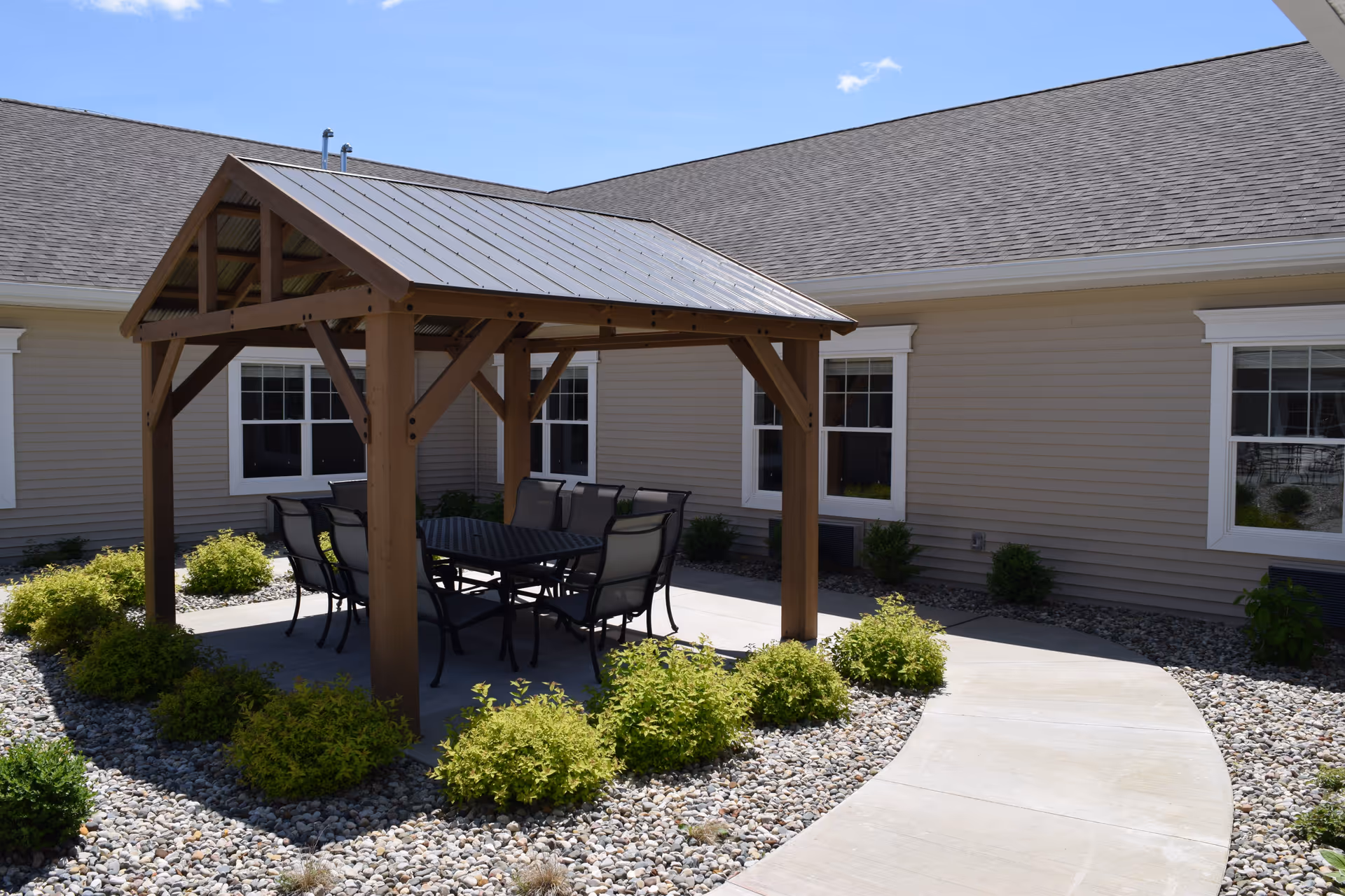 A wooden gazebo with a table and chairs on a paved courtyard surrounded by gravel, shrubs, and the exterior of the building.