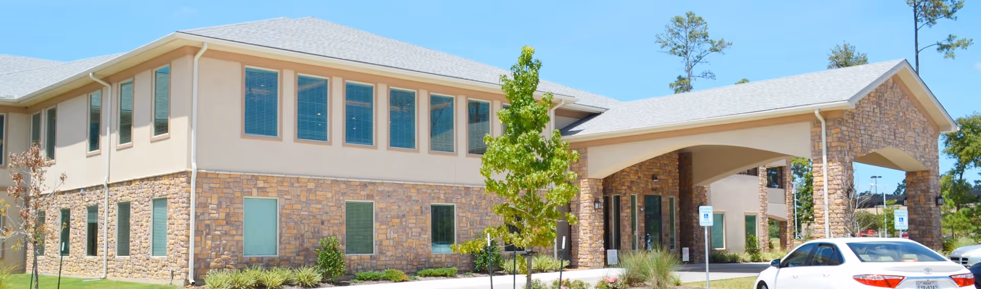 Exterior view of a two-story senior living facility building with stone and beige siding, large windows, a covered entrance, and a white car parked in front.