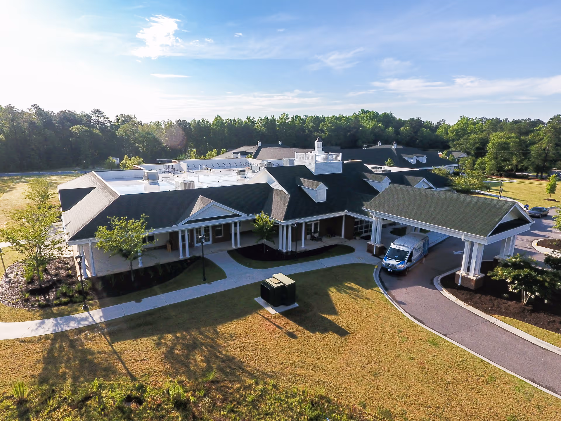 Aerial view of a single-story senior living building with a covered porte-cochere, parked van, driveway, lawn, and surrounding trees.