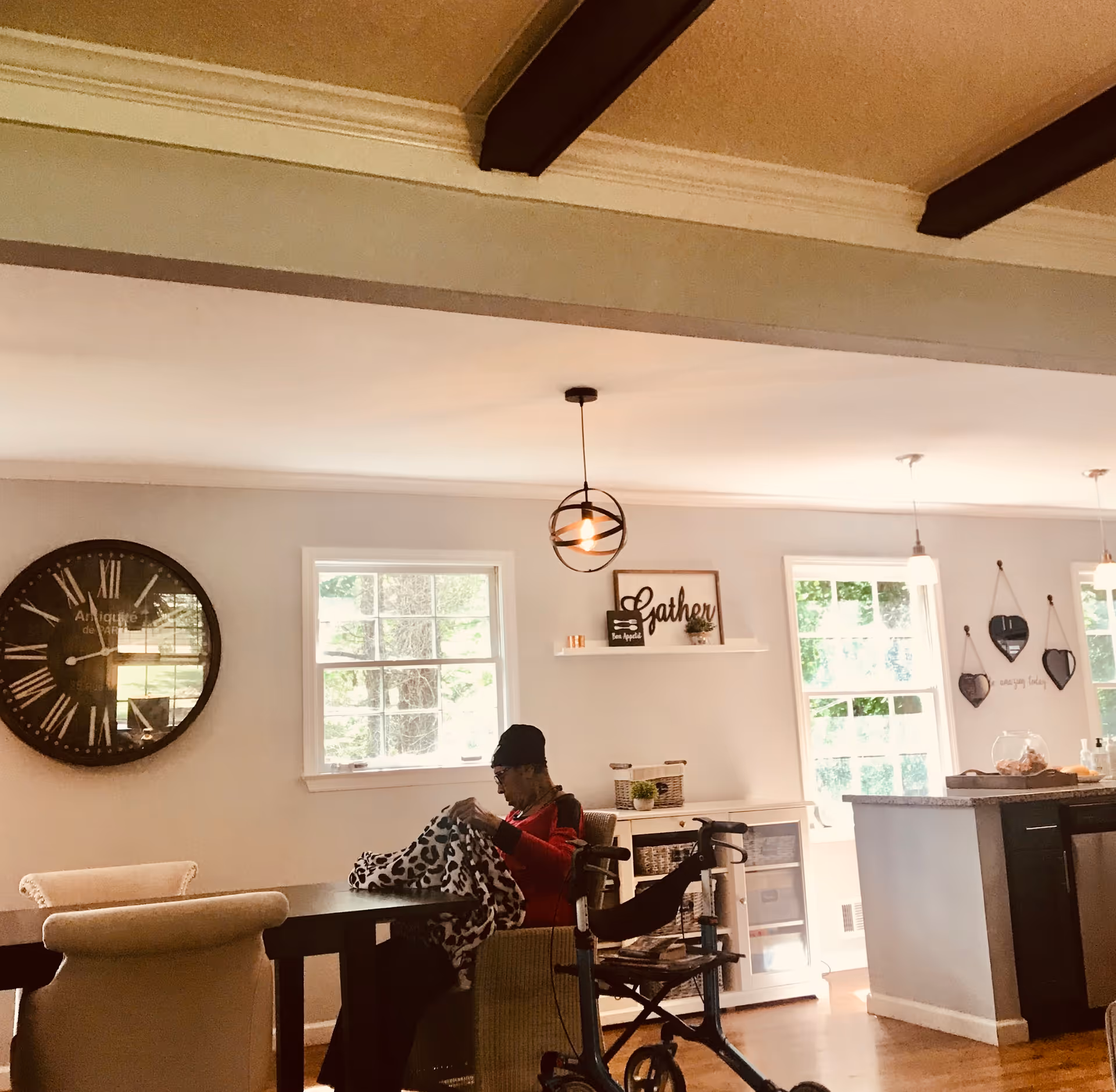 A cozy interior of a senior living facility with a woman sitting at a dining table covered with a blanket. The room features large windows letting in natural light, a large wall clock, decorative shelves with signs, and a walker nearby. The ceiling has exposed wooden beams and pendant lights hang over a kitchen island in the background.