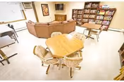 A bright common area in Kilkenny Court featuring a hexagonal wooden table with four chairs in the foreground, a brown sectional sofa facing a wall-mounted TV, and a bookshelf filled with board games and books against the wall.
