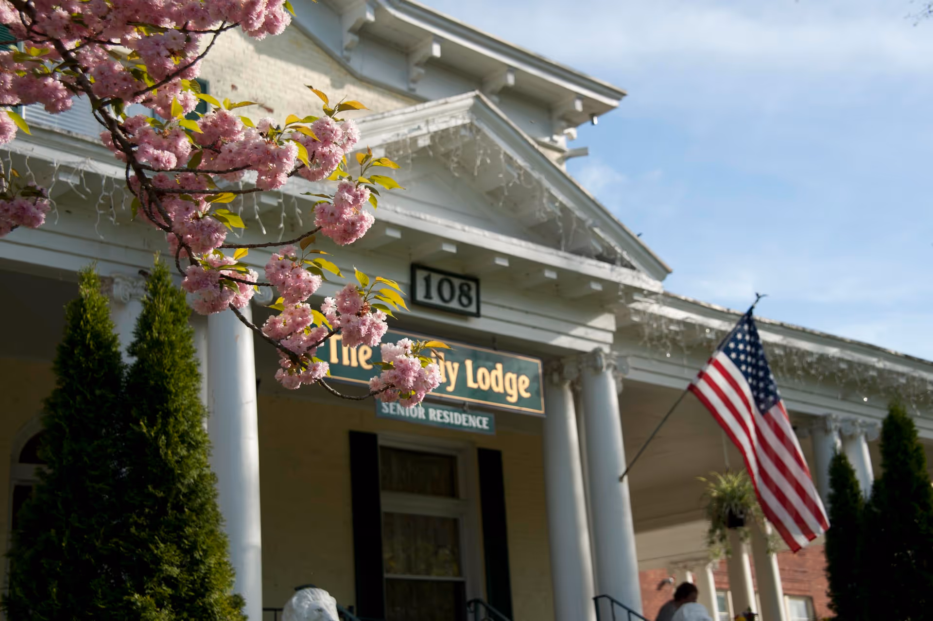 Front exterior view of Ivy Lodge Assisted Living with white columns, an American flag, and blooming pink flowers in the foreground under a clear sky.