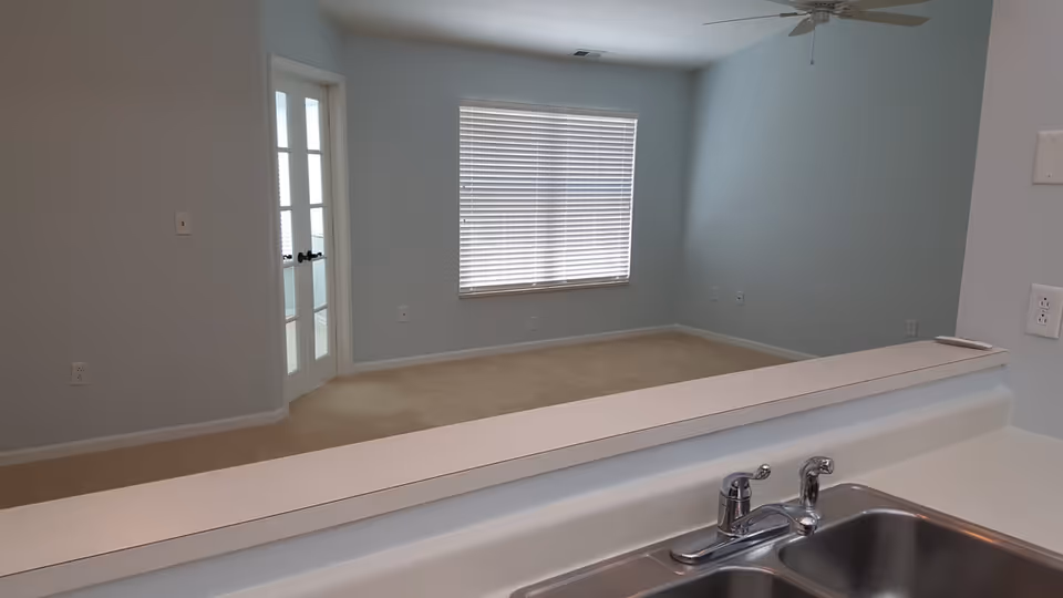 View from a kitchen sink over a pass-through counter into an empty living room with a window, French doors, and ceiling fan.