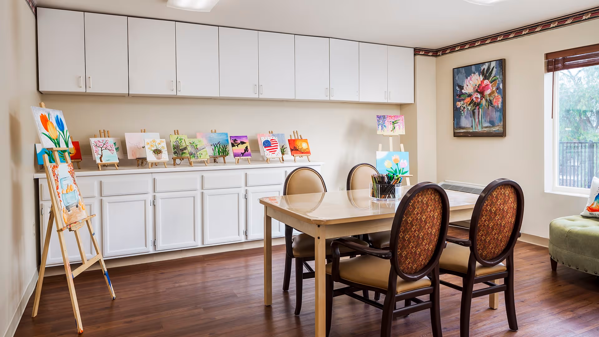 A bright art room with a wooden table surrounded by four chairs with patterned upholstery. On the table, there are containers holding paintbrushes and art supplies. Along the back wall, there is a long white cabinet with multiple small paintings displayed on easels. A larger painting of flowers hangs on the wall near a window with a view of trees outside. The room has wooden flooring and white walls.