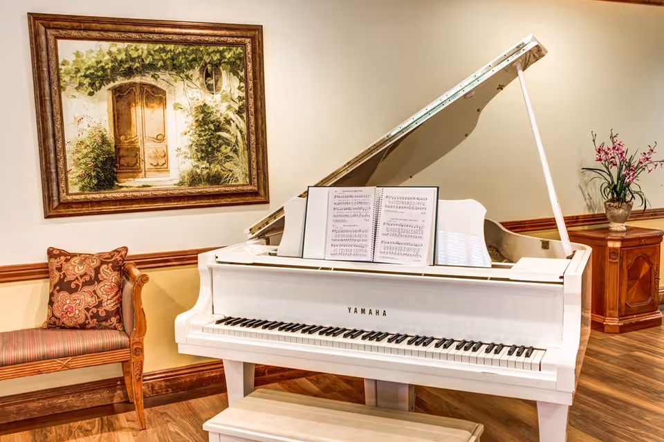 A white Yamaha grand piano with sheet music on the stand, placed in a warmly lit room with wooden flooring. To the left of the piano is a wooden bench with a patterned cushion, and above it hangs a framed painting of a wooden door surrounded by greenery. To the right, there is a wooden cabinet with a vase of pink flowers on top.